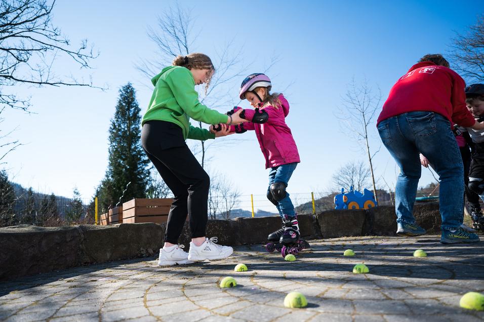 Zwei Kinder üben das Rollschuhlaufen, während eine Aufsichtsperson Unterstützung bietet; Tennisbälle liegen auf dem Boden.